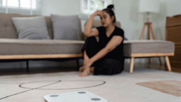 a woman sitting on her living room floor looking frustrated as she looks down at a scale