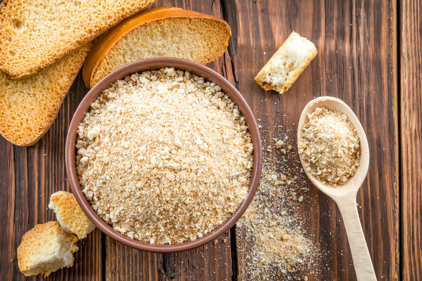 Bowl of breadcrumbs on a table next to sliced pieces of bread.