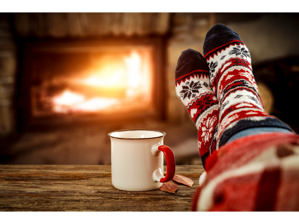 Person wearing comfy socks sitting with their feet up on a coffee table in front of a warm fire. There is a cup of tea on the table and a blanket is around their legs.  
