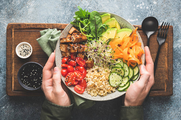 Bowl of food including tomatoes, spinach, carrots, avocados, and seasoned tofu in Male Hands On Wooden Serving Tray Background