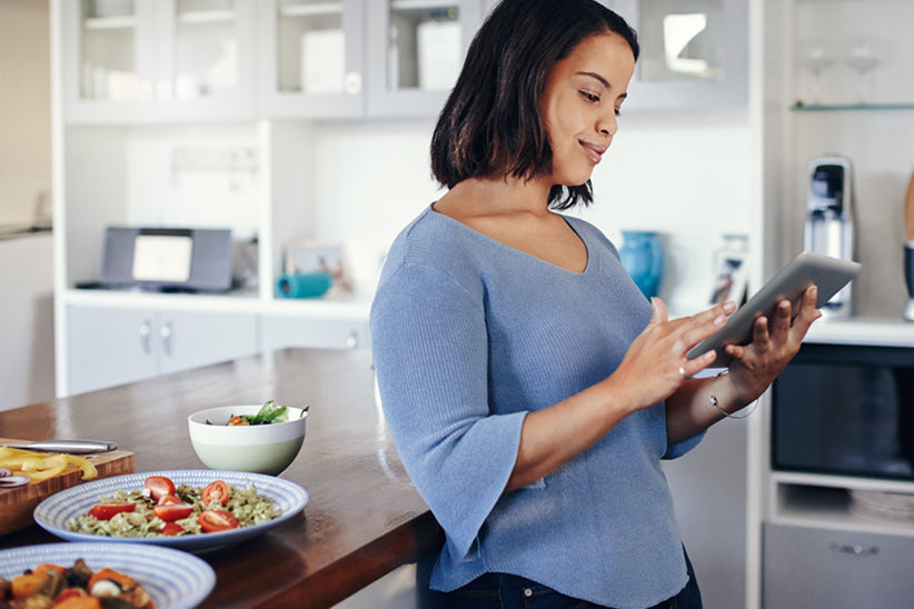woman standing in a kitchen looking on her tablet with a healthy meal on the table next to her.