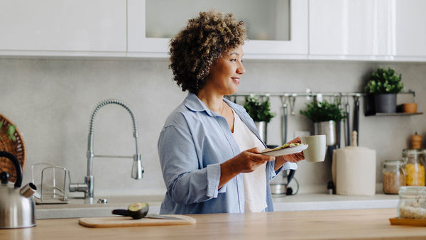 Smiling woman standing at her kitchen counter with a plate of healthy food in her hands and an avocado on the cutting board.