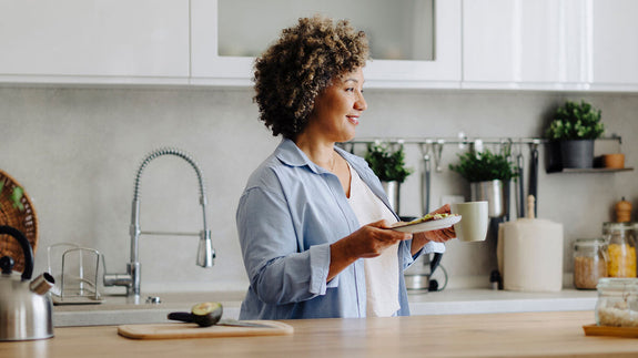 Smiling woman standing at her kitchen counter with a plate of healthy food in her hands and an avocado on the cutting board.